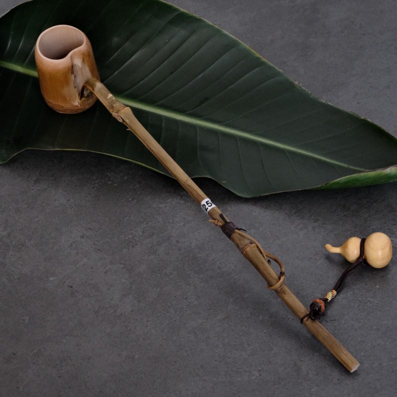 May include: A long-handled bamboo ladle with a cylindrical cup at one end and a small, light-colored gourd-shaped charm attached to the handle by a dark cord. A small white tag with the number 25 is wrapped around the center of the bamboo handle.
