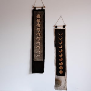 May include: Two vertical wall hangings featuring moon phase designs. The left hanging has a light-coloured panel with dark moon phases on a black background. The right hanging has a black panel with light moon phases on a light background.