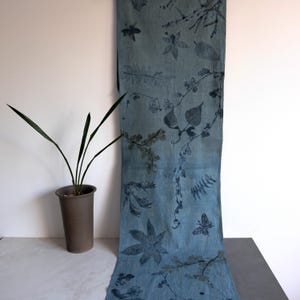 May include: A long, rectangular blue fabric runner featuring a botanical print pattern. The design includes dark blue silhouettes of various leaves, stems, flowers, and a butterfly scattered across the surface of the light blue textile material.