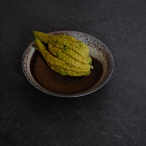 Könnte beinhalten: Eine gelbe, texturierte Buddha-Hand-Zitronenfrucht mit mehreren fingerartigen Segmenten, die in einer flachen, runden Keramikschale liegt, welche ein dunkelbraunes Inneres und einen gesprenkelten, hellen Rand mit rustikalem, verwittertem Finish aufweist.