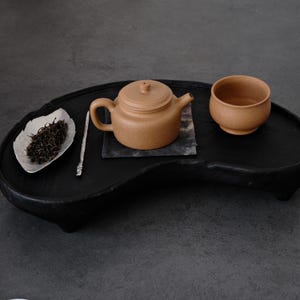 以下が含まれることがあります： 黒い木製トレイに置かれたティーセット。セットには、小さな茶色のティーポット、お揃いのティーカップ、茶葉が入った銀色の葉の形をしたお皿が含まれています。銀色の茶器がトレイの上に置かれています。