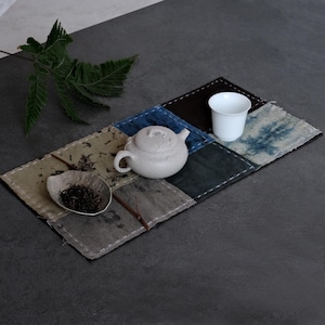 May include: A rectangular patchwork tea mat featuring various fabric squares in shades of blue, brown, and grey with visible white stitching. A light-colored ceramic teapot, a small white cup, and a shallow bowl containing loose tea leaves sit on top.