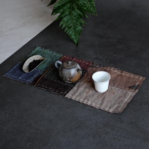 May include: A rectangular patchwork fabric tea mat featuring stitched squares in muted tones of green, blue, brown, and grey. A small ceramic teapot, a white porcelain cup, and a metal scoop containing loose tea leaves are placed on the mat.