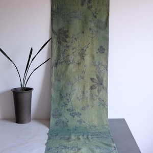 Puede incluir: Un camino de mesa textil largo y rectangular con un estampado botánico de hojas y enredaderas en verde oscuro y carbón sobre un fondo verde oliva claro. La tela muestra una textura natural y orgánica con siluetas de plantas.
