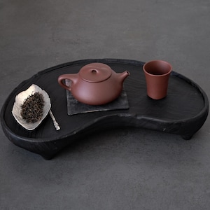 May include: A dark brown tea set on a black wooden tray. The set includes a small teapot, a cup, and a silver leaf-shaped dish with tea leaves. The tray has a unique, organic shape.