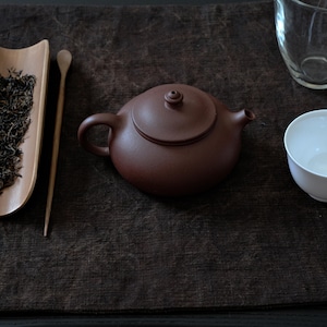 May include: A brown ceramic teapot with a rounded body, small spout, lid, and handle. A wooden scoop and a tray of loose tea leaves are to the left. A white teacup and a clear glass are to the right. The items are on a dark brown surface.