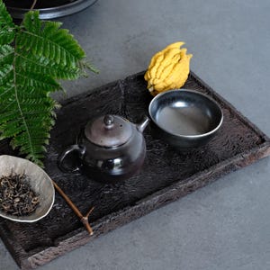 May include: A rectangular dark brown wooden tray with a textured surface holds a small dark brown teapot, a matching dark bowl, a light-colored dish containing loose tea leaves, and a bright yellow Buddha's hand citron fruit.