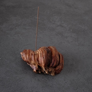 Peut inclure: Un porte-encens rustique en bois naturel, de couleur brun rougeâtre avec des veines apparentes. Un fin bâton d'encens cuivré est inséré dans le support. Le support est posé sur une surface gris foncé. Le bois présente un aspect texturé.