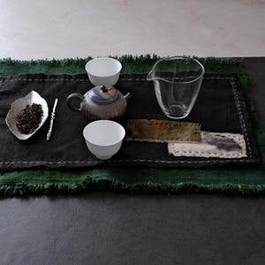 Puede incluir: Un juego de té con una tetera de cerámica oscura, dos tazas de porcelana blanca, una jarra de cristal transparente, un plato en forma de hoja con té a granel y un agitador de metal, todo dispuesto sobre un tapete de tela oscura con bordes deshilachados.