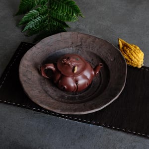 Peut inclure: Une théière en argile brun foncé avec une surface texturée et organique, dotée d'un couvercle aux petits détails sculptés, posée au centre d'un bol en céramique assorti, large et peu profond, au bord irrégulier et fini mat.