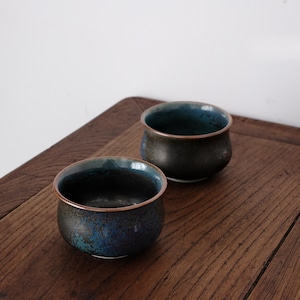 Könnte beinhalten: Zwei Keramikschalen mit dunkelblauer und brauner Glasur. Die Schalen haben eine runde Form mit einem leicht ausgestellten Rand. Das Innere der Schalen ist tiefblau, das Äußere ist gesprenkelt braun und blau. Die Schalen stehen auf einer Holzoberfläche.