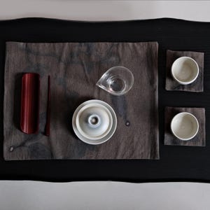 May include: A white ceramic mug with a black handle and the text "But First, Coffee" in a bold, black font. The mug is filled with a dark liquid, likely coffee, and sits on a light-colored surface.