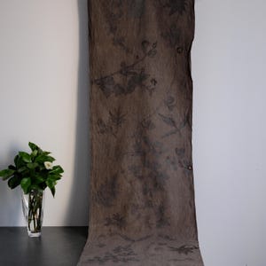 May include: A long, rectangular piece of brown fabric featuring a dark, botanical print of leaves and branches scattered across its surface. The textile has a textured, slightly translucent appearance with raw, uneven edges along the sides.