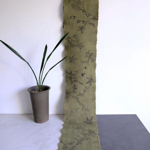 May include: A long, narrow strip of olive green fabric featuring a dark botanical print of various leaves and stems. The fabric has raw, deckled edges and is displayed vertically, showcasing a repeating pattern of natural foliage in a dark gray tone.