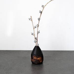 Puede incluir: Un jarrón decorativo con la parte superior negra y una base marrón texturizada. El jarrón contiene ramas con adornos plateados en forma de hoja. El arreglo se encuentra sobre un fondo blanco.
