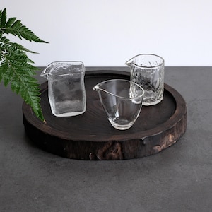 Puede incluir: Tres recipientes de vidrio transparente con superficies texturizadas y picos vertedores dispuestos sobre una bandeja circular de madera oscura. Los recipientes varían en forma, con un diseño rectangular y dos redondeados.