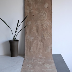Puede incluir: Una pieza larga y rectangular de tela color canela con un estampado botánico de siluetas blancas de hojas y plantas. La tela muestra un tono natural y terroso con una textura visible y bordes deshilachados, exhibiendo varios patrones.