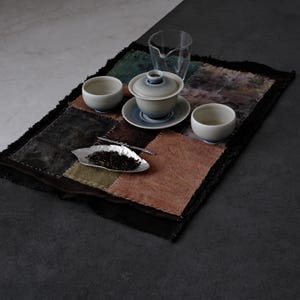 Może przedstawiać: Zestaw do herbaty z przykrytym gaiwanem, dwiema małymi ceramicznymi czarkami i przezroczystym szklanym dzbankiem na prostokątnej, wielokolorowej macie z tkaniny patchworkowej z postrzępionymi brzegami. Srebrzyste naczynie w kształcie liścia zawiera herbatę.