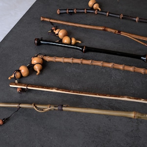 May include: A collection of five long, slender bamboo walking sticks or staffs, each featuring distinct natural textures, varying shades of brown and black, and decorative small, light-colored gourd ornaments attached with dark cord.