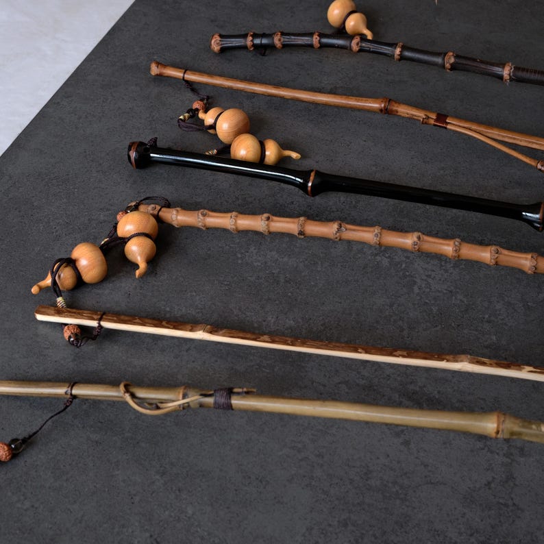 May include: A collection of five long, slender bamboo walking sticks or staffs, each featuring distinct natural textures, varying shades of brown and black, and decorative small, light-colored gourd ornaments attached with dark cord.