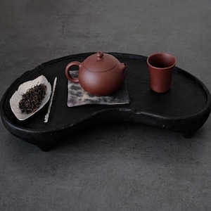 May include: A dark brown tea set on a black wooden tray. The set includes a small teapot, a teacup, a silver leaf-shaped dish with tea leaves, and a tea utensil. The tray has a unique, organic shape.
