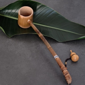 May include: A bamboo water ladle with a cylindrical cup and a long, slender handle. The handle features a small white sticker with the number 258 and a black cord attached to a small, light-colored wooden gourd-shaped charm at the end.