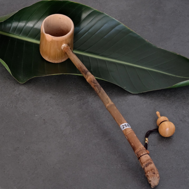 May include: A bamboo water ladle with a cylindrical cup and a long, slender handle. The handle features a small white sticker with the number 258 and a black cord attached to a small, light-colored wooden gourd-shaped charm at the end.