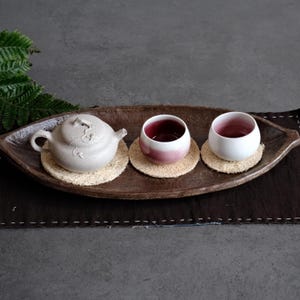 Op de afbeelding: Een lichtgrijze keramische theepot met een gestructureerd wijnrankreliëf staat op een ronde beige onderzetter, vergezeld door twee kleine wit-roze gradiënt keramische kopjes op bijpassende onderzetters, op een lange, bruine keramische schaal.
