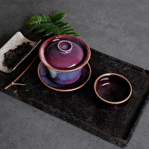 Könnte beinhalten: Ein Keramik-Teeservice bestehend aus einer Gaiwan-Schale mit Deckel und einer passenden Tasse, beide mit einer tiefvioletten und blauen gesprenkelten Glasur und braunen Rändern. Dazu gehört eine kleine weiße Schale mit losem Tee und ein Teelöffel.