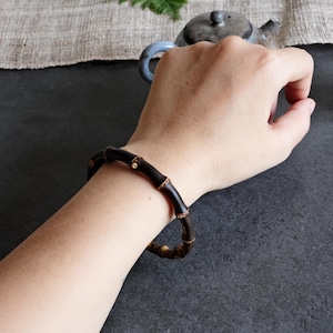 Könnte beinhalten: Ein dunkelbraunes Armband, das einem segmentierten Bambusstab nachempfunden ist. Das kreisförmige Accessoire weist natürlich wirkende Knoten und Rillen entlang seiner Länge auf, mit einer glatten, polierten Oberfläche, die die tiefen Erdtöne betont.