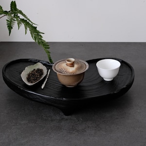 Puede incluir: Una bandeja de té negra en forma de corazón contiene un juego de té de cerámica. El juego incluye un cuenco de té con tapa, una taza blanca pequeña y un plato en forma de hoja con hojas de té. Una ramita de helecho verde añade un toque natural.