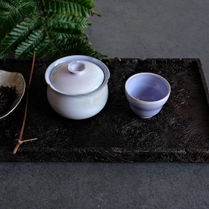 Könnte beinhalten: Ein helles, lavendelfarbenes Keramik-Teeservice, bestehend aus einer Schale mit Deckel und einer passenden Tasse. Die Schale hat einen kleinen blumenförmigen Knauf auf dem Deckel. Das Set ist mit einer kleinen Schale mit Teeblättern und einem Holzstab arrangiert.