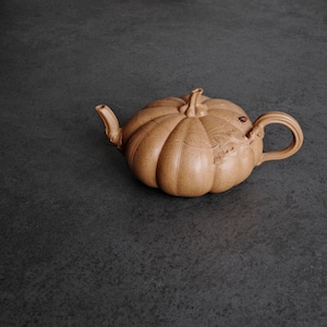 May include: A light brown, textured clay teapot shaped like a pumpkin with a curved spout, a central stem-like lid handle, and a looped handle. The surface features vertical ridges and a small, dark red accent near the handle attachment point.