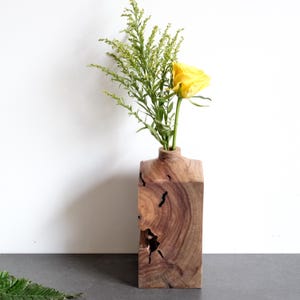 Puede incluir: Un florero rectangular de madera con veta natural y un nudo oscuro prominente en la parte frontal. El florero contiene una rosa amarilla brillante y una rama alta de follaje verde, creando un contraste entre la madera y las flores.