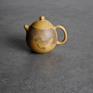 May include: A round, matte yellow teapot featuring a dark, intricate line drawing of a dragon wrapping around its body. The teapot has a small, centered lid knob, a curved handle, and a short spout, all crafted from a smooth, uniform material.