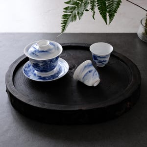 Könnte beinhalten: Ein Set aus blau-weißen Porzellan-Teetassen und einer Teeschale mit Deckel und passender Untertasse, alle mit Landschaftsdesign. Die Artikel sind auf einem dunklen, runden Holztablett angeordnet. Ein Farnzweig ist im Hintergrund zu sehen.