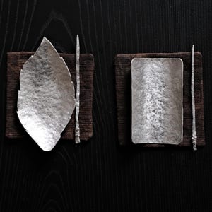 May include: Two silver-colored serving pieces, one leaf-shaped and one rectangular, rest on dark brown square mats. Each has a thin, silver-colored utensil. The items are set against a dark wood grain background, creating a minimalist aesthetic.