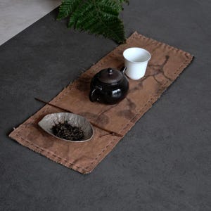 May include: A rectangular brown fabric tea mat with visible stitched edges, featuring a black ceramic teapot, a white ceramic cup, a small textured grey dish containing loose tea leaves, and a thin wooden stick placed across the mat surface.