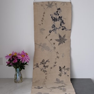 Op de afbeelding: Beige textiel ophanging met een botanische print van bladeren en wijnranken in grijstinten. Het textiel is opgehangen aan een metalen ring. Een vaas met roze bloemen staat links.