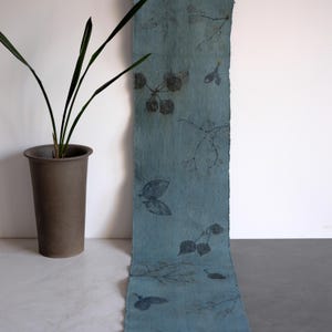 May include: A long, rectangular strip of blue fabric featuring a dark botanical print of leaves and twigs. The fabric has a textured, fibrous appearance with raw, deckled edges. Beside the fabric stands a tall, matte brown cylindrical vase.