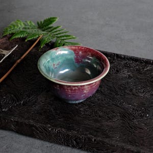 May include: A small ceramic bowl featuring a glossy glaze with blended shades of deep magenta, purple, and teal. The bowl has a smooth, rounded rim and a slightly tapered base, showcasing a reactive finish that creates unique color transitions.
