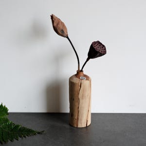 Puede incluir: Un florero cilíndrico de madera clara con una grieta vertical en el costado y una pequeña etiqueta blanca marcada con el número 025 cerca del cuello. Dos vainas de loto secas de color marrón sobre tallos largos, finos y oscuros están dentro.