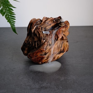 May include: A natural brown wood incense burner with a textured, gnarled surface and a small white tag labelled 393. A lit incense cone sits on top, releasing a thin stream of white smoke that flows downward over the wood grain onto the surface.