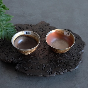 May include: Two small, round ceramic bowls with metallic rims. The left bowl has a dark brown interior and a textured, mottled brown exterior with a label reading 126. The right bowl has a light tan interior and a similar exterior with a label reading 127.