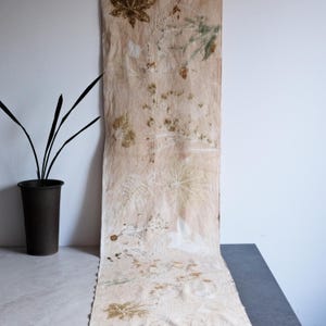 Puede incluir: Una pieza larga y rectangular de tela de color beige claro con estampados botánicos en tonos marrón, bronceado y verde apagado. El textil muestra varios patrones de hojas y plantas impresos en su superficie, creando un diseño natural.