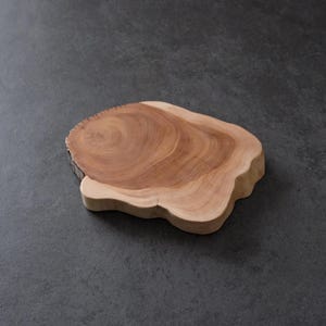 May include: A natural wood cutting board with a unique, organic shape. The board features a light brown color with darker wood grain patterns, and a live edge. The board is approximately 12 inches long and 9 inches wide.