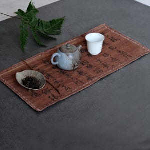 May include: A rectangular brown fabric mat featuring black calligraphy, a small grey ceramic teapot, a white ceramic cup, a leaf-shaped dish containing loose tea leaves, and a green fern frond placed on a dark grey surface.