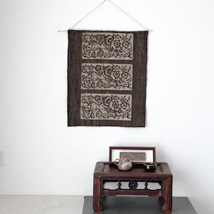 May include: A dark brown wall hanging with three rectangular panels featuring floral designs. The wall hanging is suspended from a wooden dowel with a string. Below, a dark wooden table displays a teapot, cup, and framed artwork.