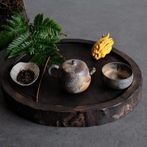 Puede incluir: Una bandeja redonda de madera marrón oscuro contiene una tetera de cerámica con esmalte gris y bronceado moteado, un cuenco de cerámica a juego, un plato pequeño irregular con hojas de té sueltas, una hoja de helecho verde y un fruto mano de Buda amarillo.
