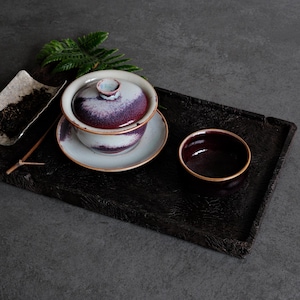 Könnte beinhalten: Ein Gaiwan-Teeservice aus Keramik mit Deckel und Untertasse, das eine weiße Basis mit einer tiefvioletten und blauen Sprenkelglasur aufweist. Daneben stehen eine passende kleine Teeschale und eine kleine Schale mit losen Teeblättern auf einem dunklen Tablett.